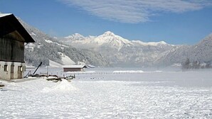Een houten hut met een met sneeuw bedekte dak, een klein tuinhuisje en een steiger die uitkomt op een bevroren meer, omringd door besneeuwde bergen.
