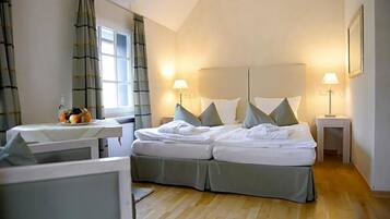 A hotel room with a bed, two bedside lamps, a window with curtains, a small table with a fruit bowl, and a framed picture on the wall.