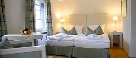 A hotel room with a bed, two bedside lamps, a window with curtains, a small table with a fruit bowl, and a framed picture on the wall.