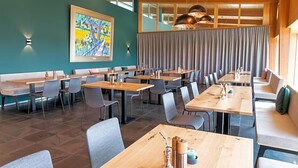Een moderne eetruimte met meerdere tafels en stoelen, een groot schilderij aan de muur en uitzicht op het buitenlands gebied door grote ramen.