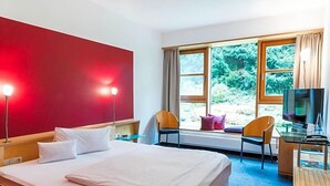 Hotelkamer met een bed, bureau, televisie en raam met uitzicht op groen.