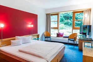 Quarto de hotel com cama, escrivaninha, televisão e janela com vista para o verde.
