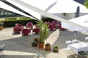 Uma área externa com assentos vermelhos e um guarda-sol branco, cercada por plantas em vasos.