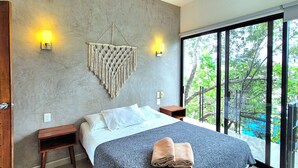 A modern bedroom with a large bed, two bedside tables, a ceiling fan, and a view of greenery through large windows. - Mi Tulum (Tulum)