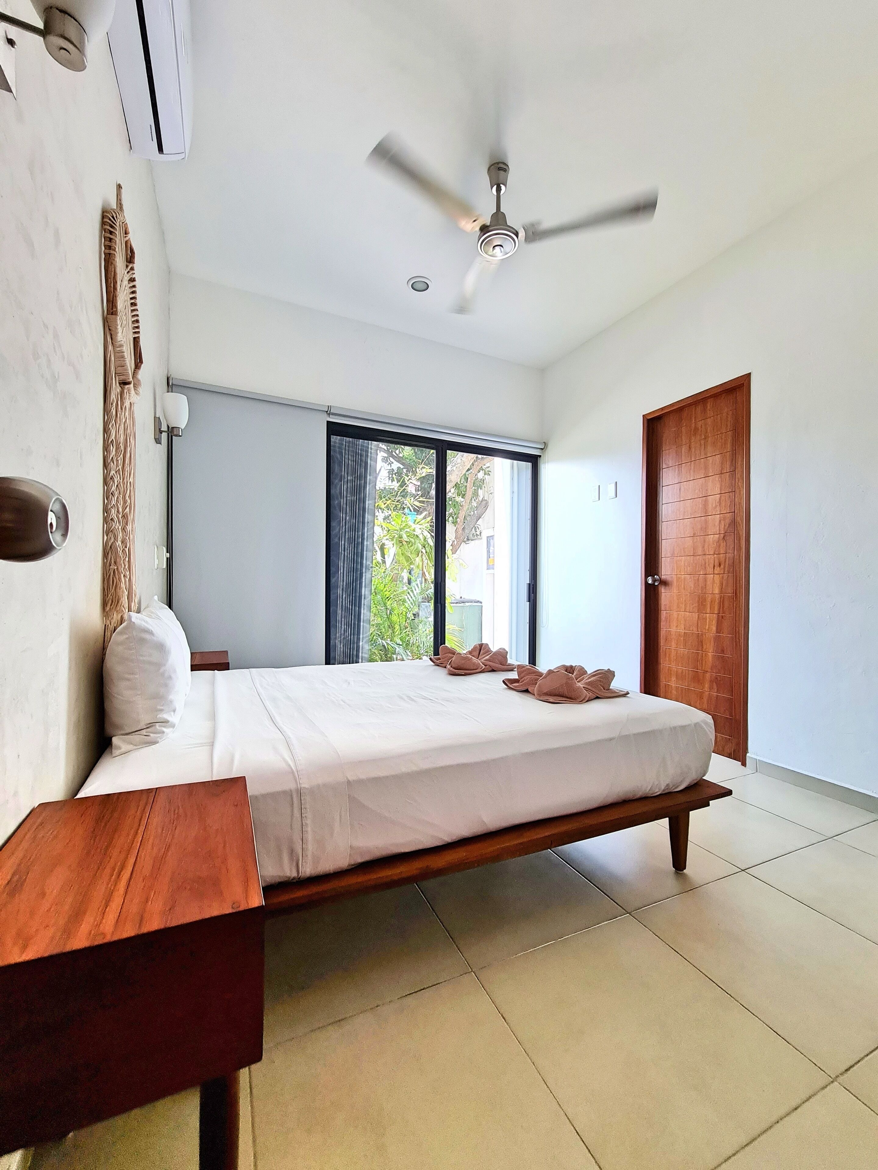 A bedroom with a bed, a ceiling fan, a window with curtains, and a wooden door.