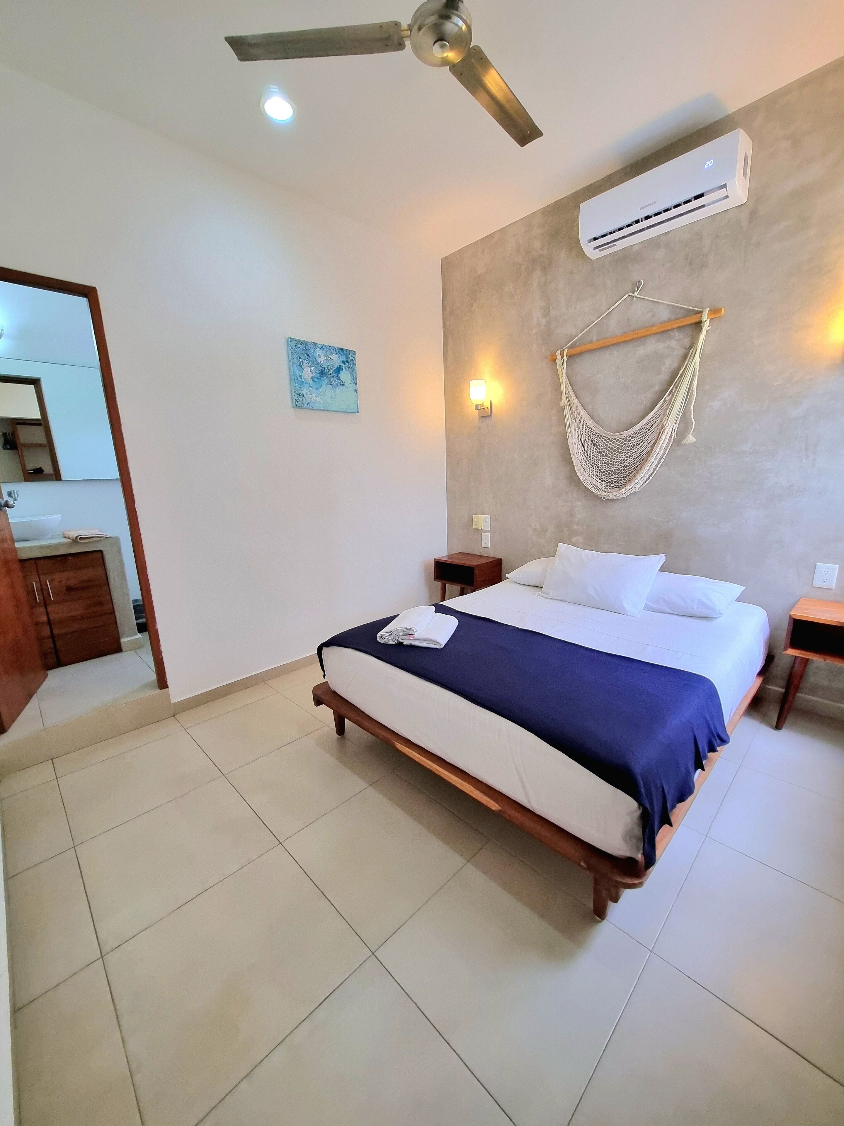 A bedroom with a bed, a ceiling fan, an air conditioner, a hammock, a nightstand, and a bathroom visible in the background.