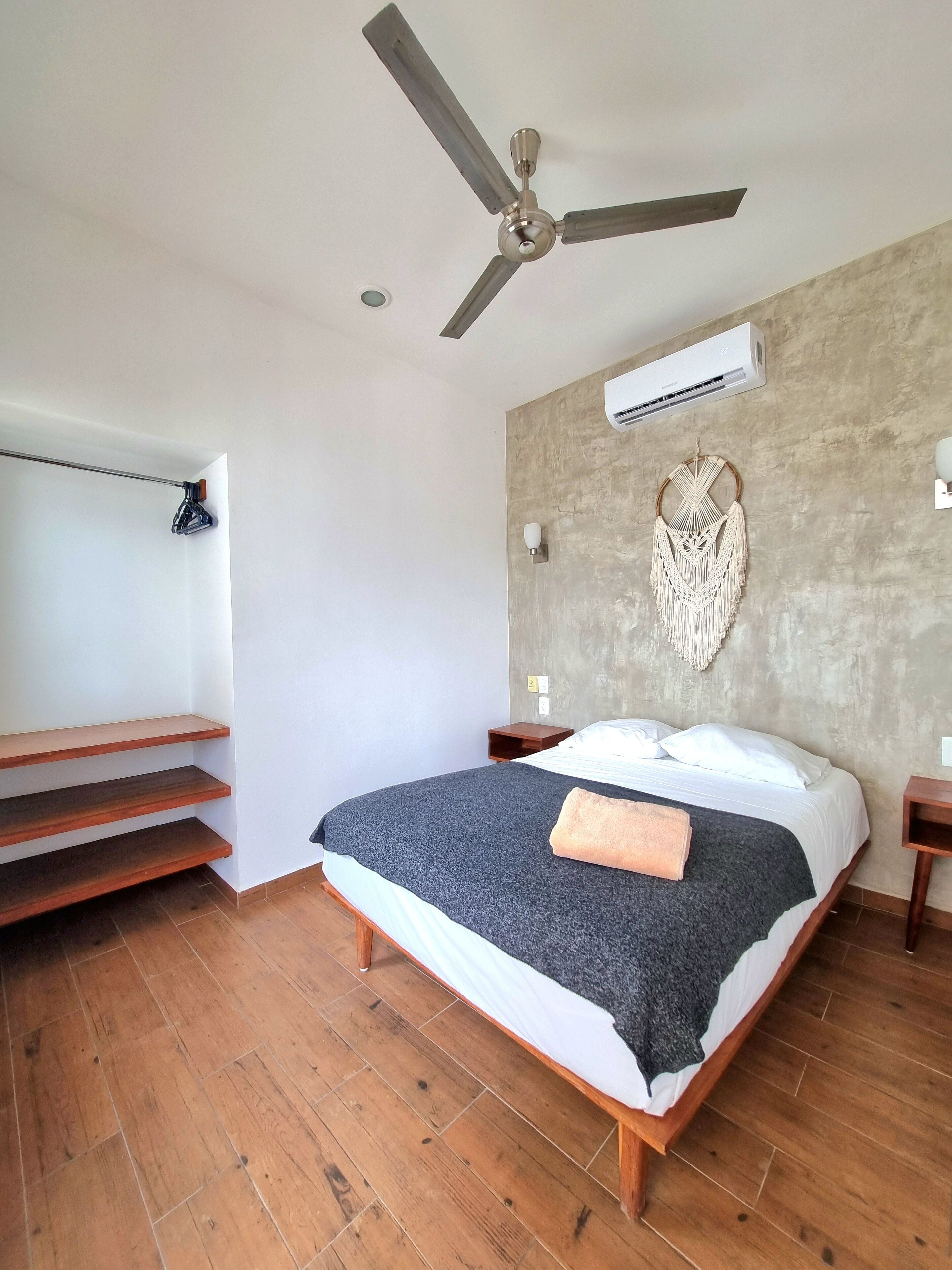 Un dormitorio con cama, suelo de madera, ventilador de techo, unidad de aire acondicionado, tapiz de macramé y una escalera.