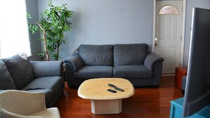 Una sala de estar con un sofá gris, una mesita de centro color beige, un sillón blanco y un ventanal con vista a un árbol.