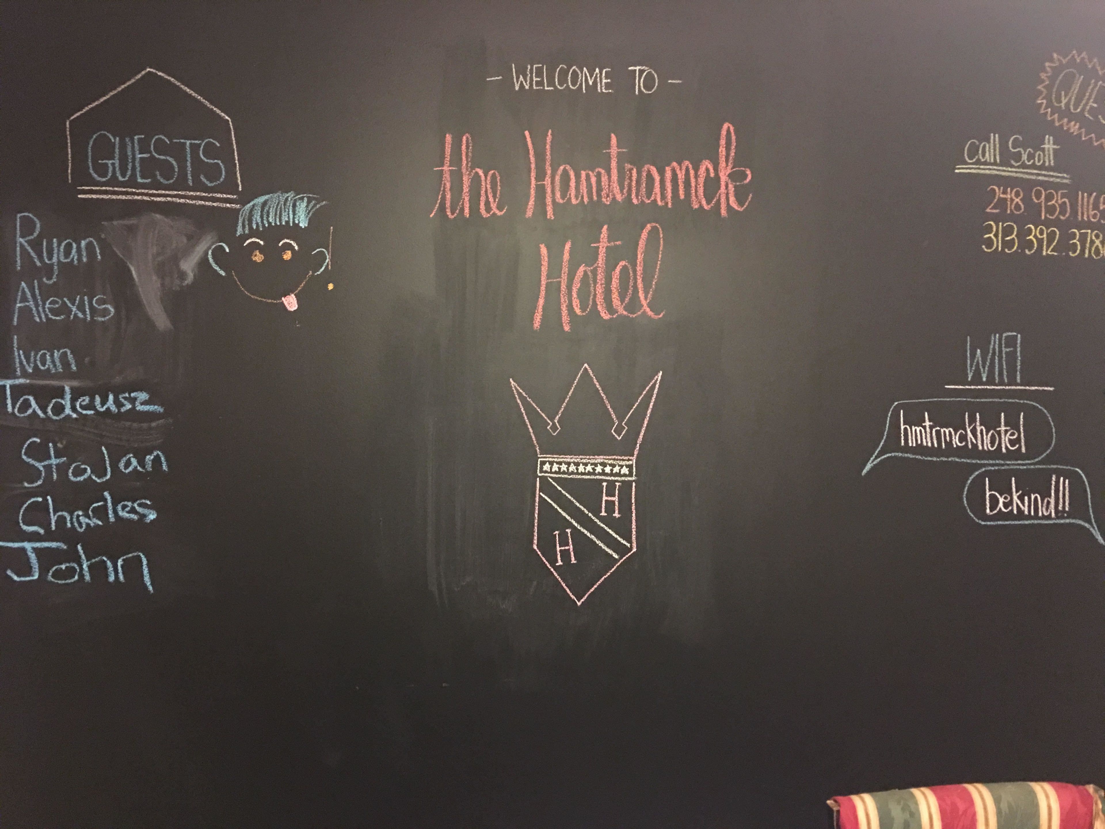 Una pizarra con un mensaje de bienvenida al 'Hamtramck Hotel', una lista de huéspedes e información de contacto, incluyendo un número de teléfono y detalles de WiFi.