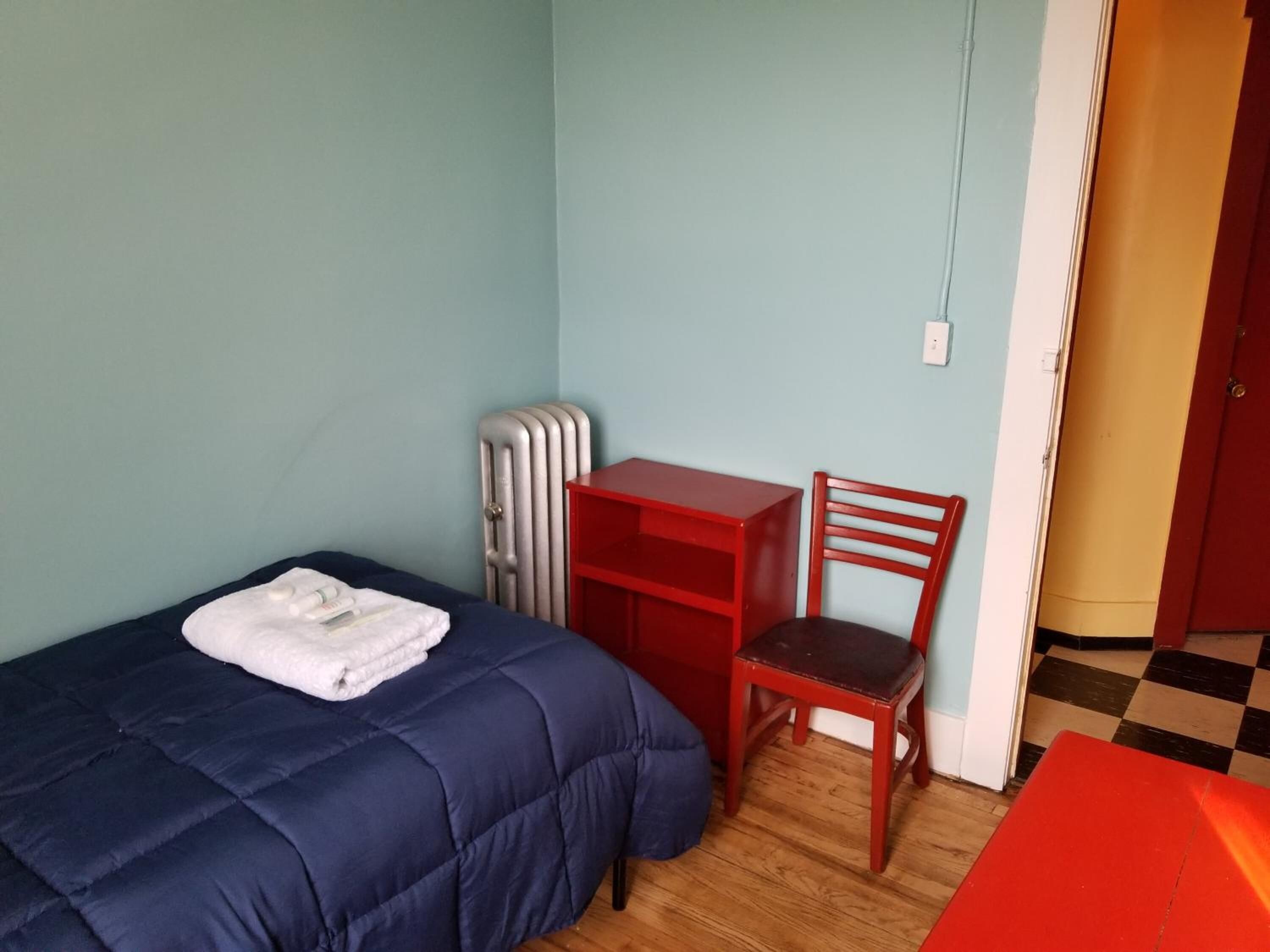 A small room with a bed, a red desk, a chair, a radiator, and a door.