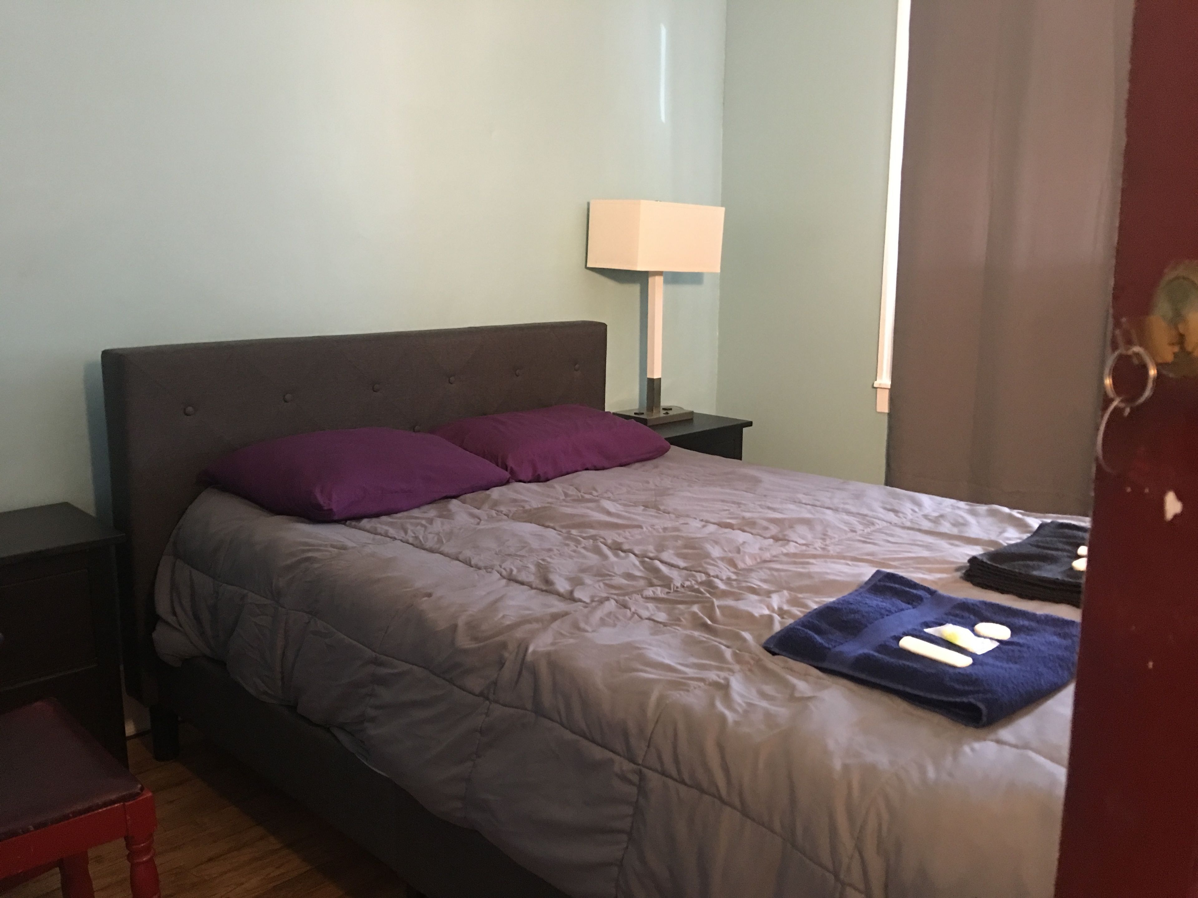 Quarto com cama, duas almofadas, mesa de cabeceira, abajur e porta.