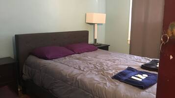 Quarto com cama, duas almofadas, mesa de cabeceira, abajur e porta.