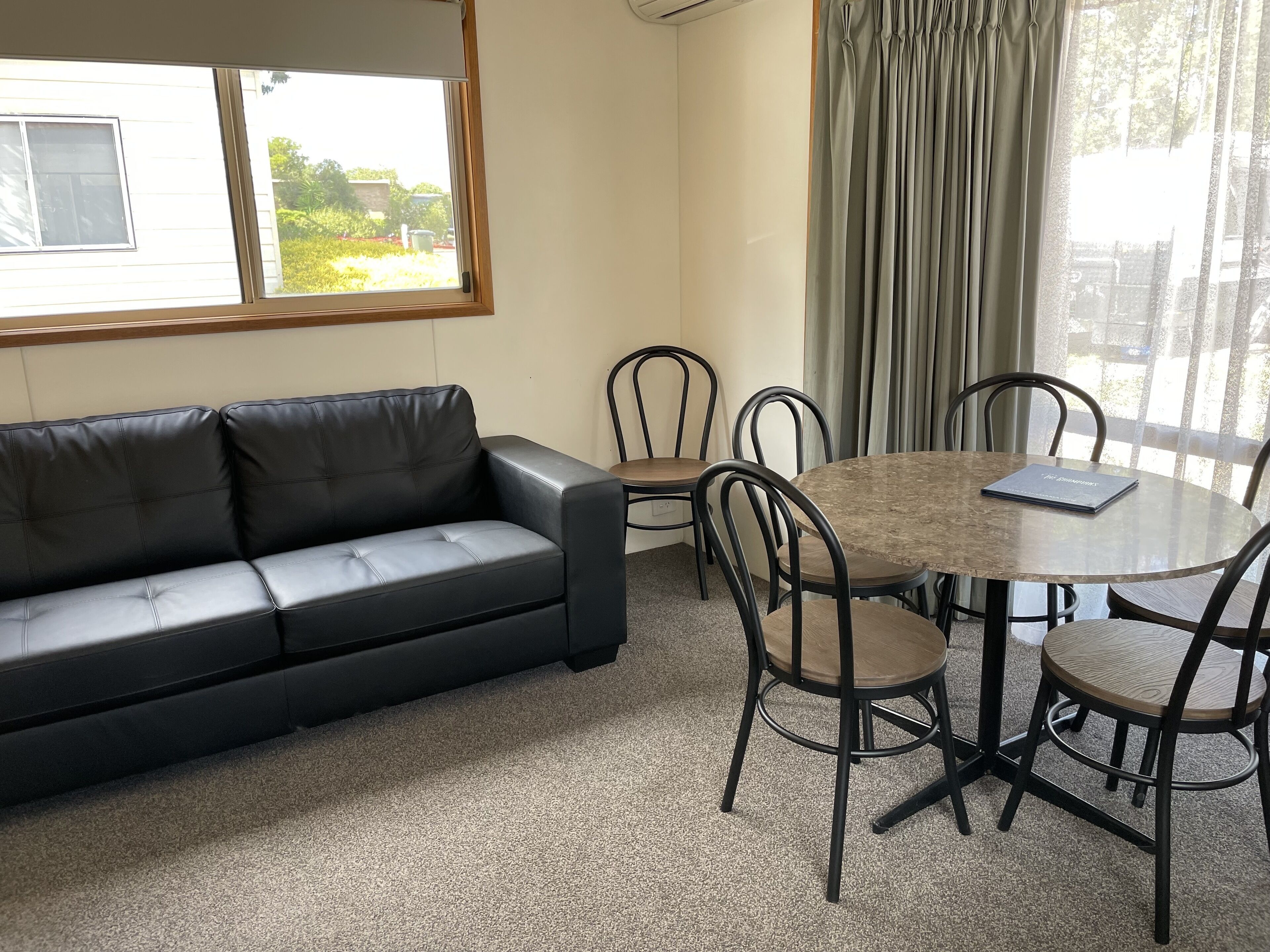 A room with a black leather sofa, a round wooden table with four chairs, a window with curtains, and a view of greenery outside.