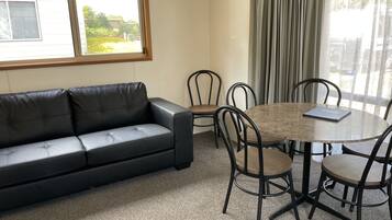 A room with a black leather sofa, a round wooden table with four chairs, a window with curtains, and a view of greenery outside.