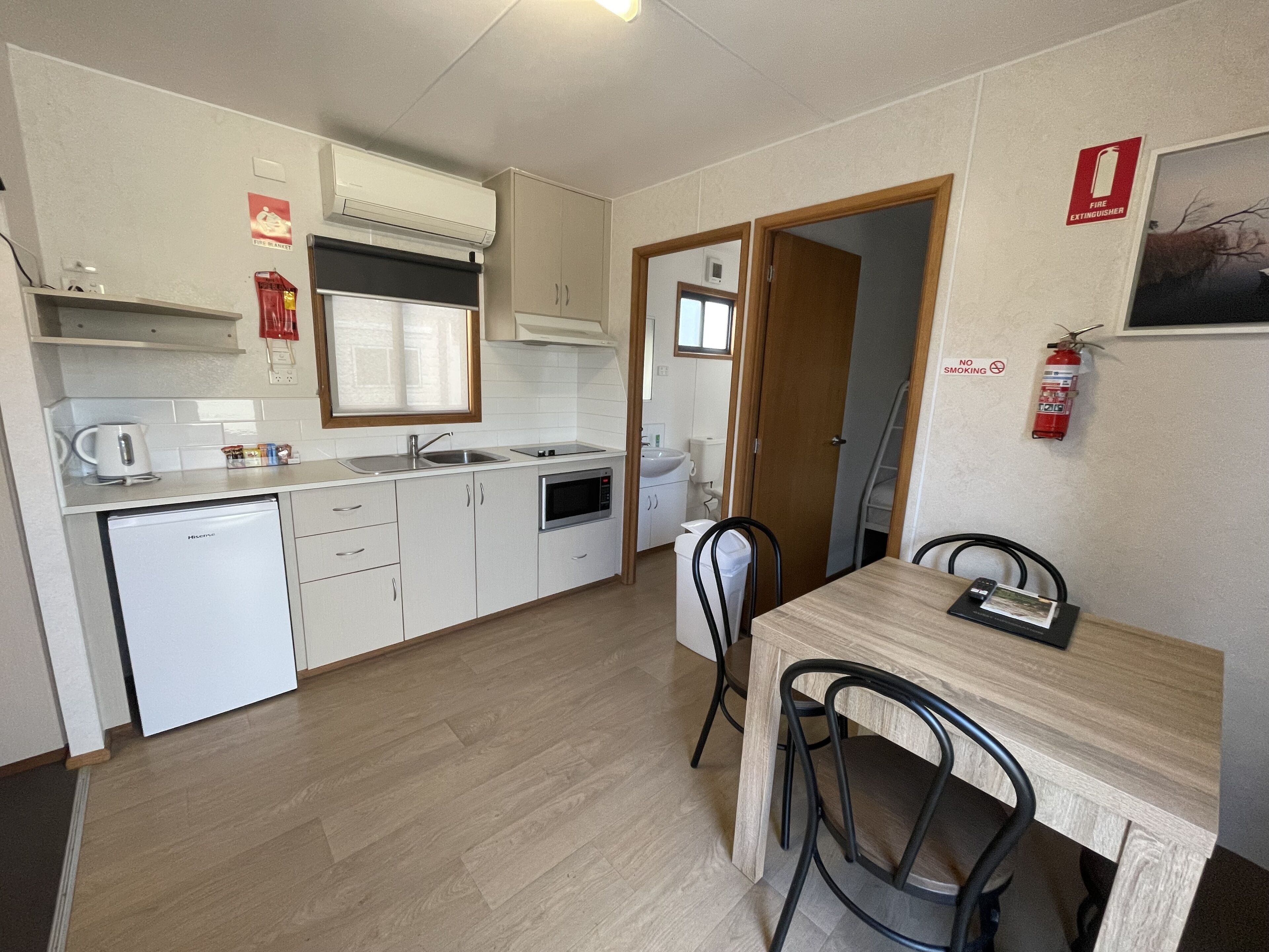 A compact kitchen with white cabinets, a built-in oven, and a microwave. There is a dining table with two chairs and a fire extinguisher mounted on the wall.