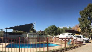 A fenced swimming pool area with a shaded structure, a fenced parking area, and a tall tree in the background.