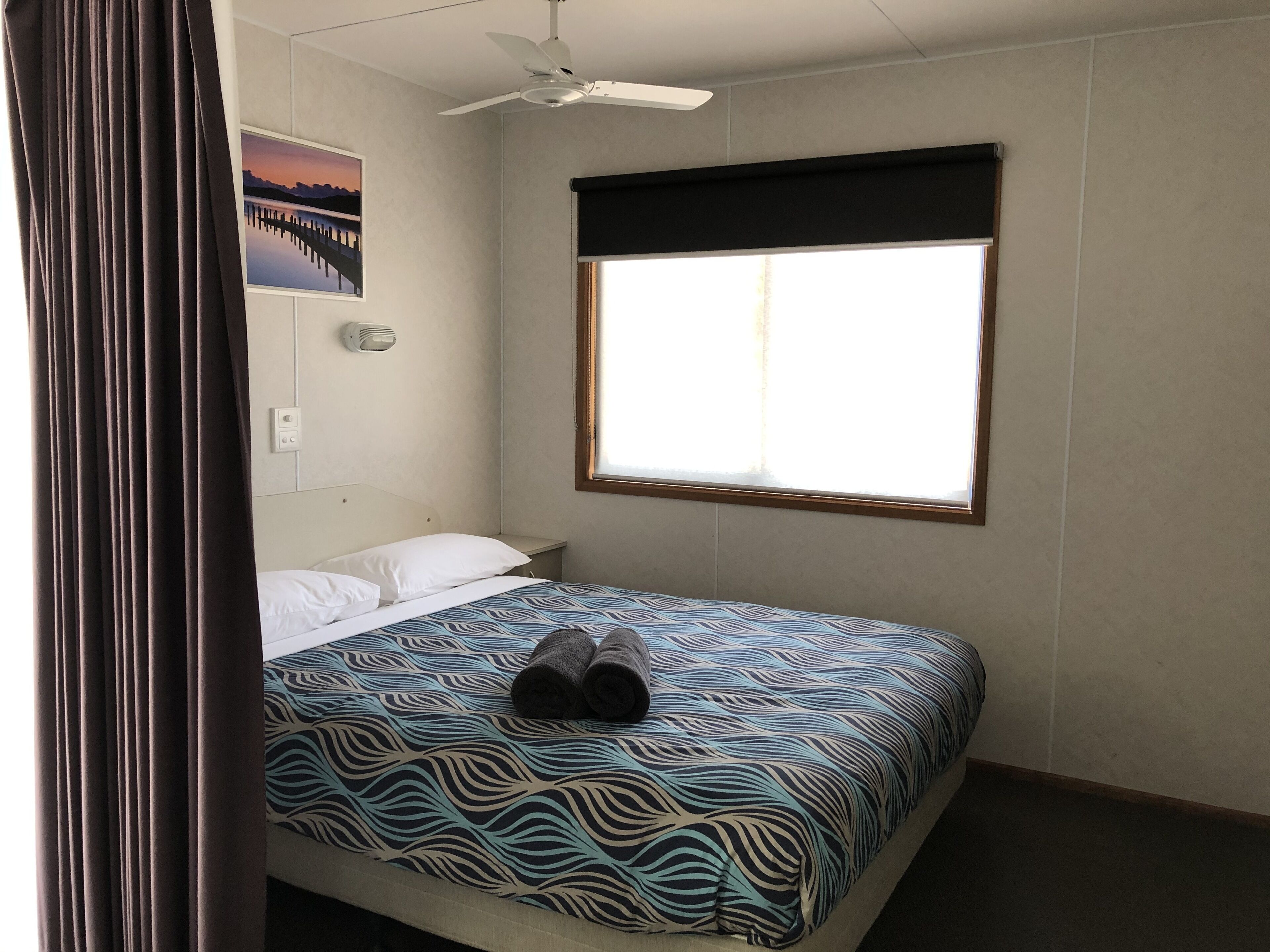 A hotel room with a bed, a ceiling fan, a window with blinds, a wall-mounted light, and a painting.