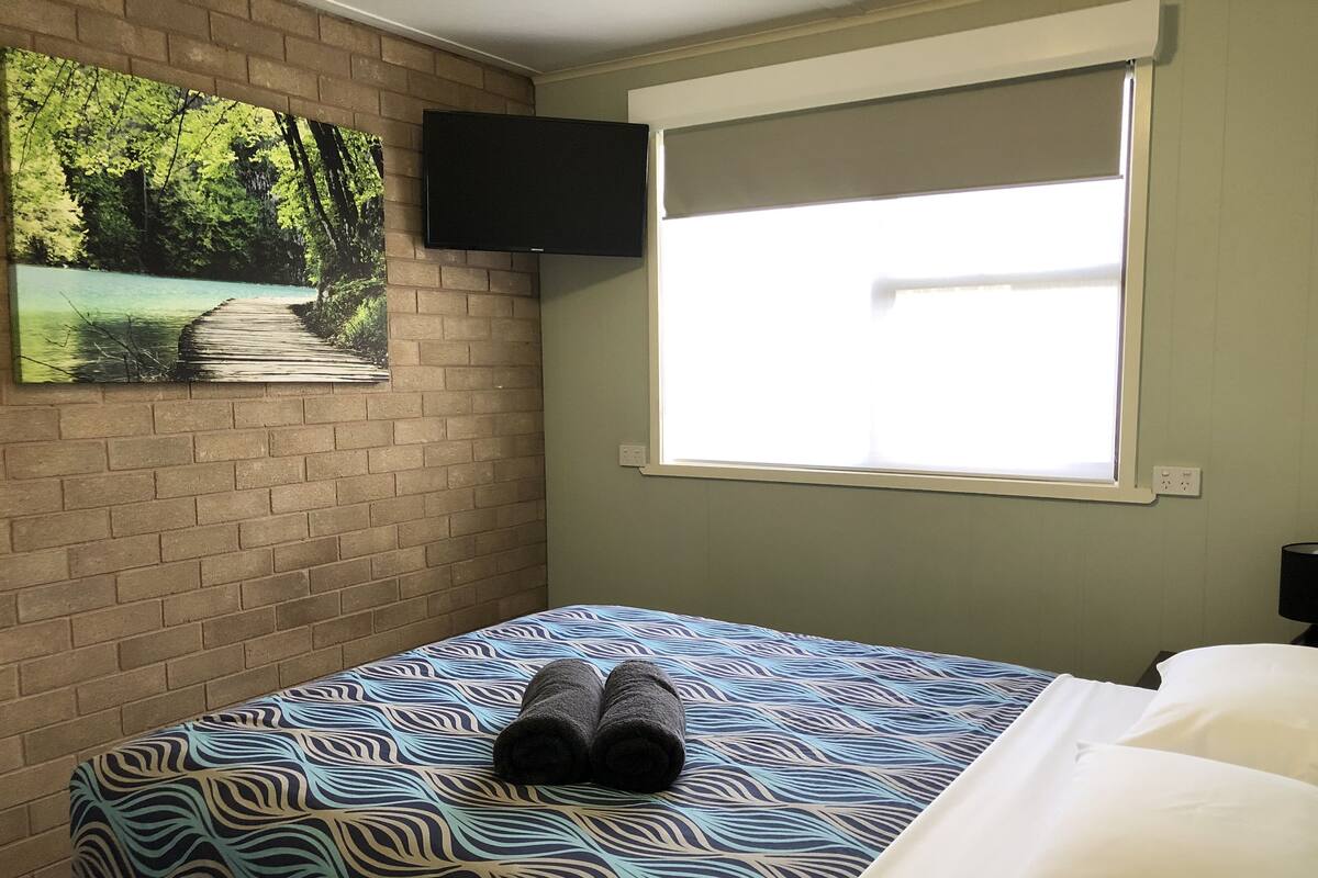 Ein Zimmer mit einem Bett, einem Fenster, einem an der Wand befestigten Fernseher und einem Waldgemälde.
