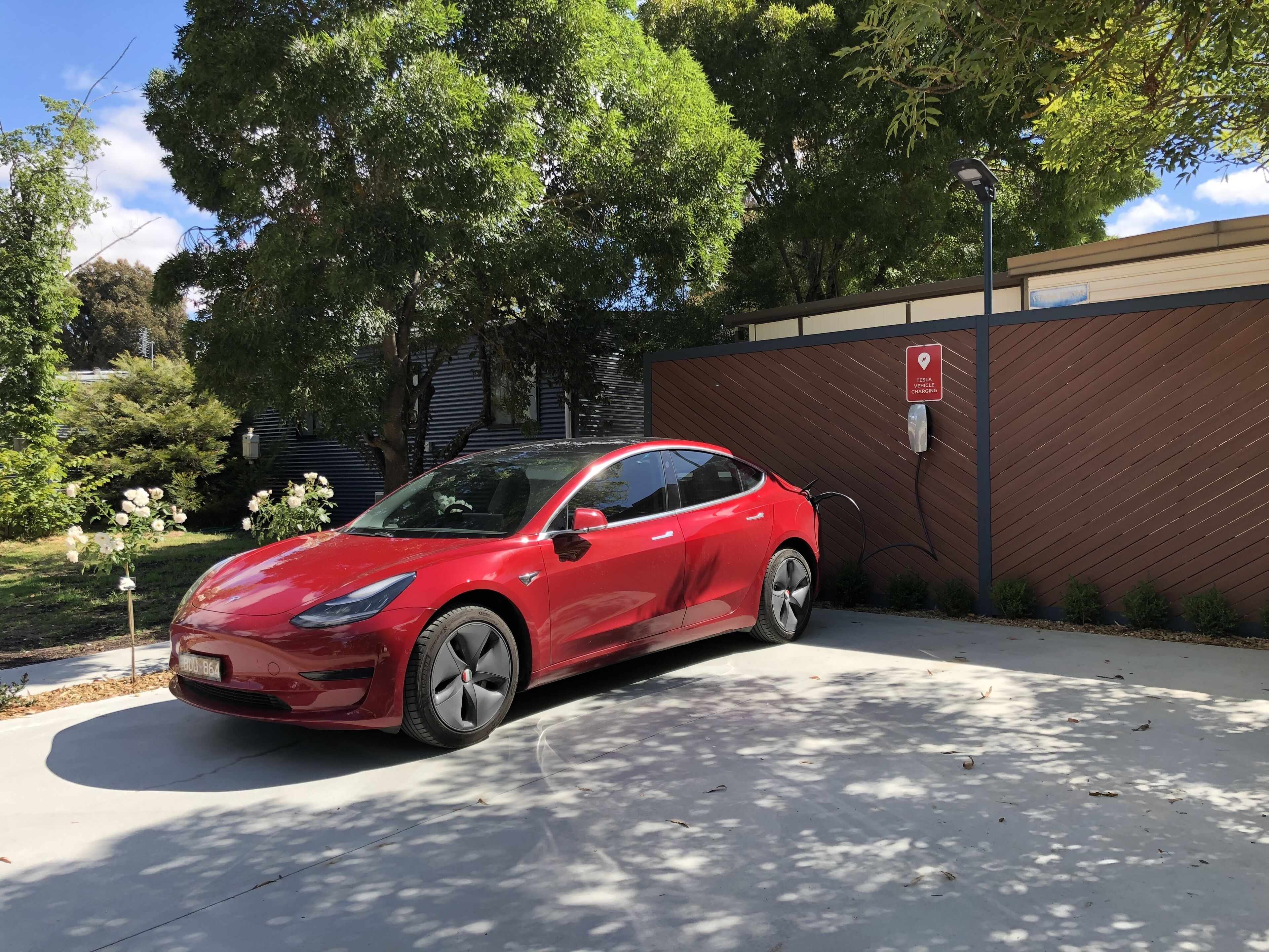 A red Tesla charging in a covered parking area with trees and a building in the background.