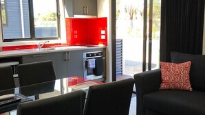 A modern living room with a black sofa and a red patterned pillow, a glass dining table with chairs, a kitchen area with a red countertop, and a large window offering a view of the outdoors. - Acacia Caravan Park (Ararat)