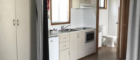 Cocina compacta con fregadero, estufa y microondas. Hay una ventana pequeña con persianas y una puerta que da al baño.