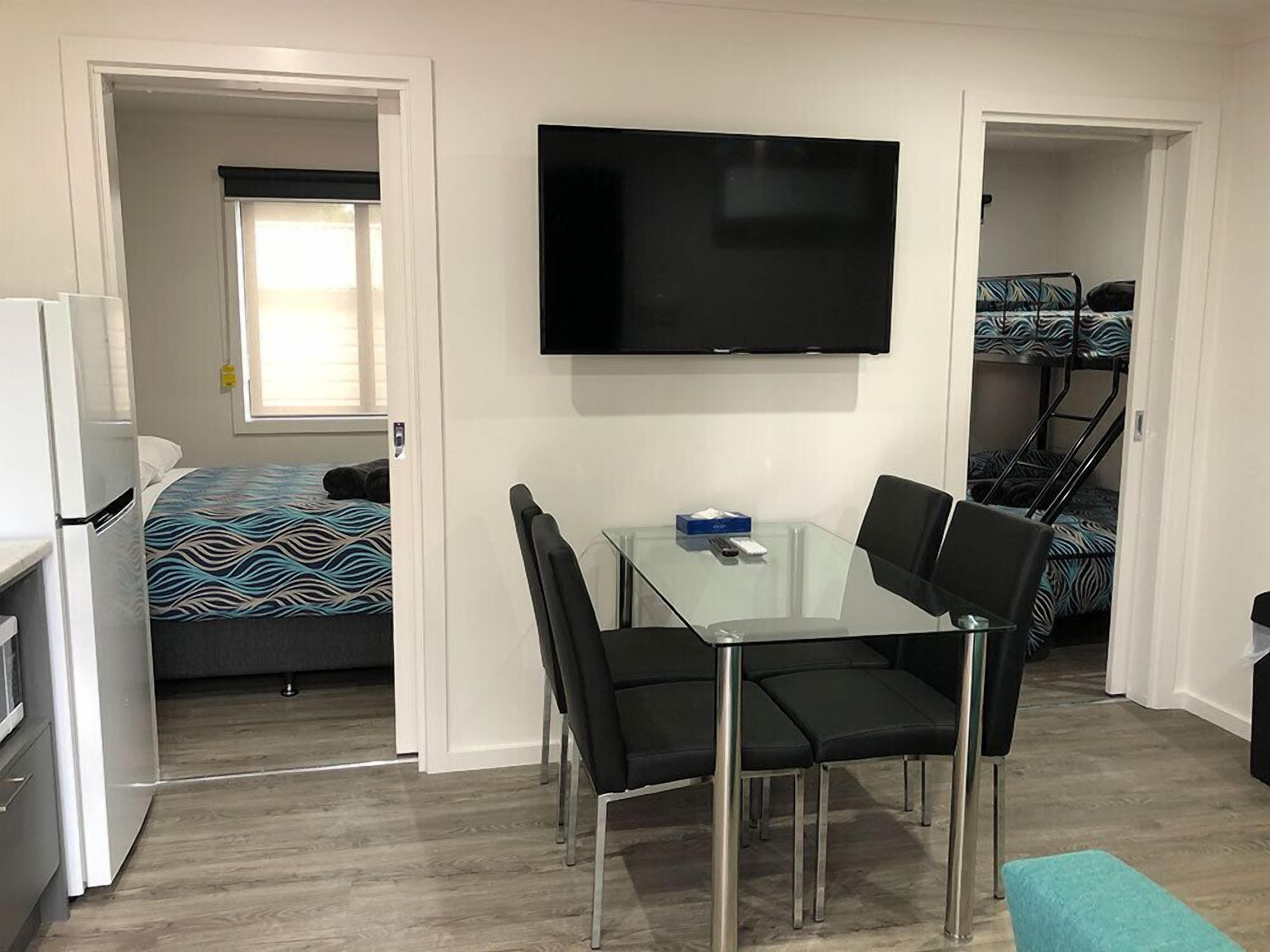 Habitación compacta con cama, televisor montado en la pared, pequeña mesa con sillas y zona de kitchenette.