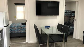 Habitación compacta con cama, televisor montado en la pared, pequeña mesa con sillas y zona de kitchenette.