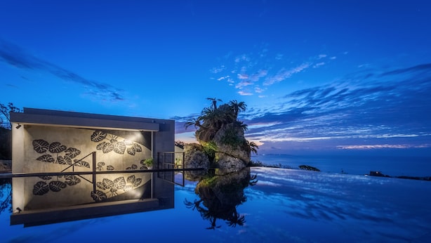 A modern building with a reflective pool, palm trees, and a sunset sky. - Hanalee Villa Kouri (Nakijin)