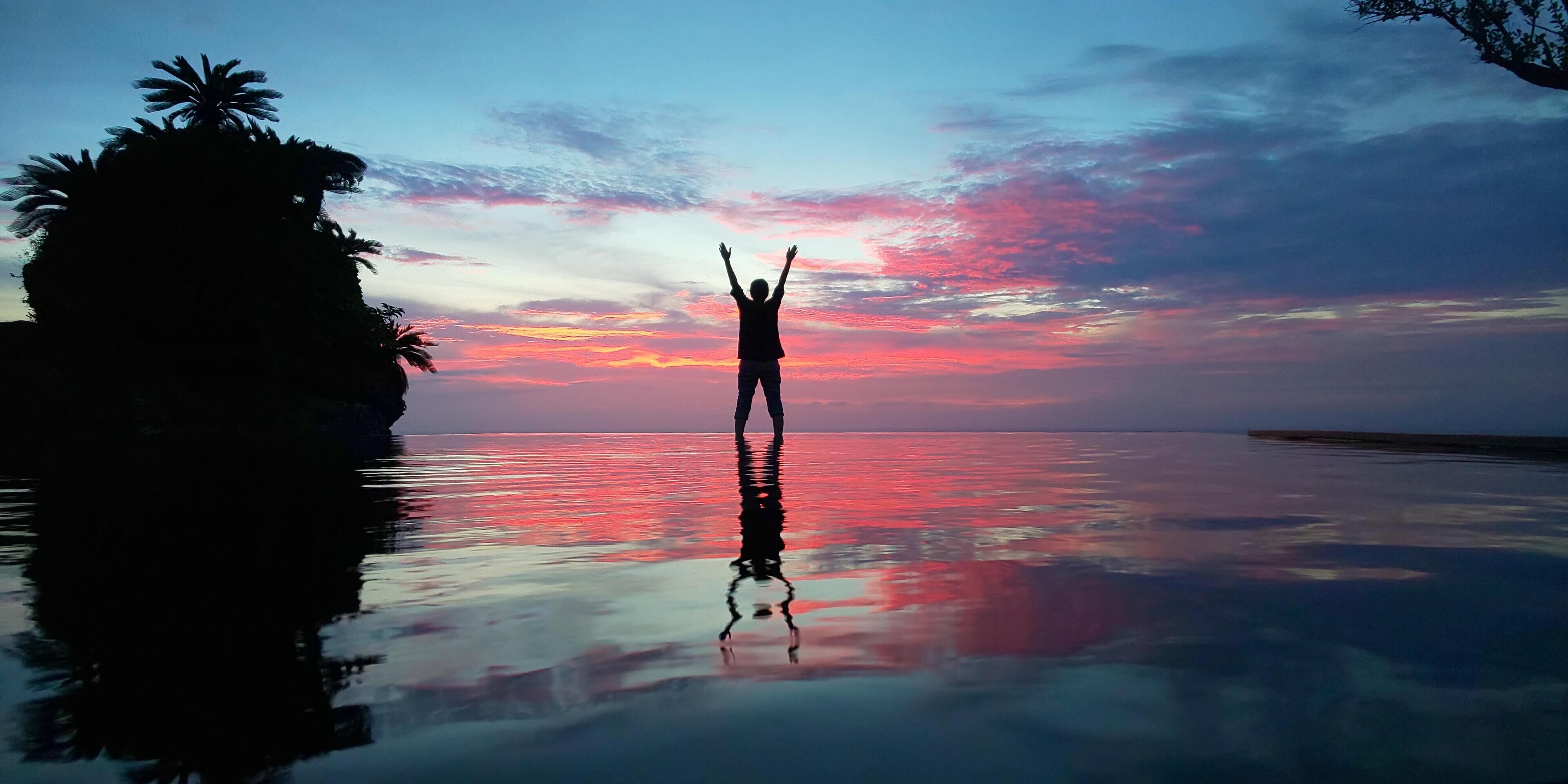 La silhouette d’une personne, les bras levés, se détache sur le ciel d’un coucher de soleil surplombant une étendue d’eau calme, avec des palmiers sur la gauche.