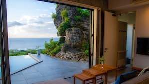 A modern living room with a large sliding glass door leading to a balcony overlooking a rocky cliff and the sea. - Hanalee Villa Kouri (Nakijin)