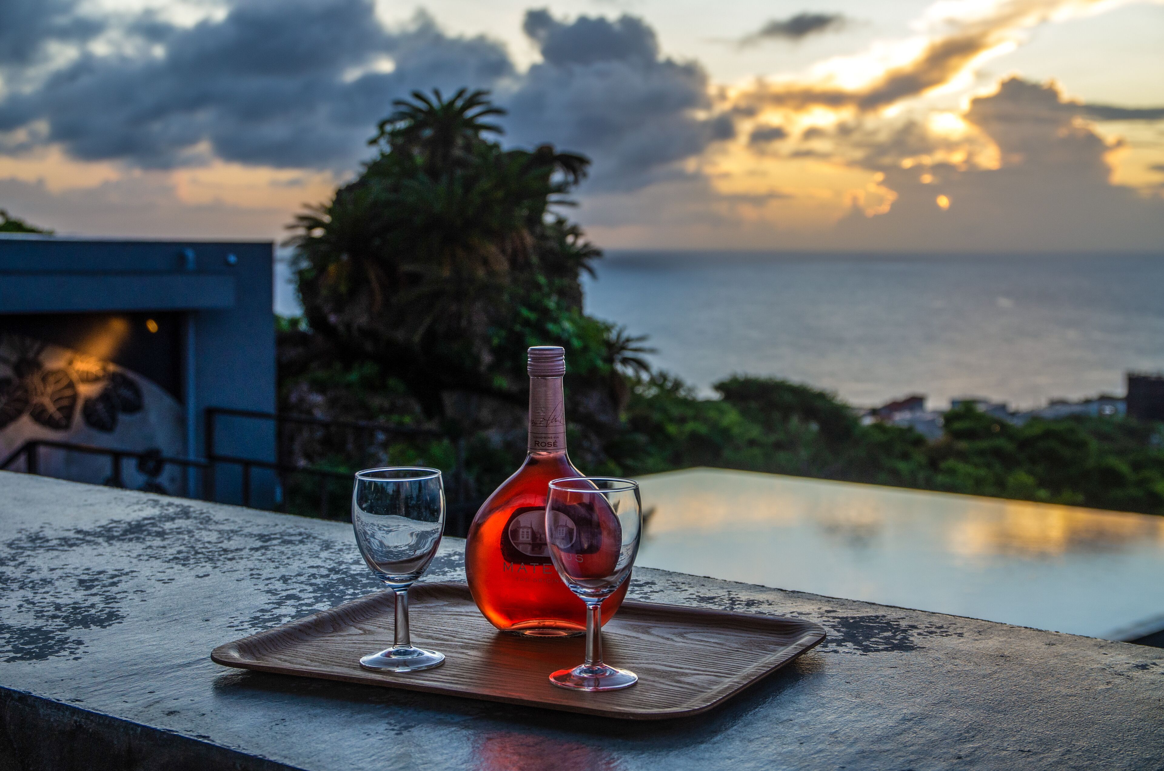 Une bouteille de vin rosé et deux verres sur un plateau en bois, près d’une piscine avec vue sur l’océan et les palmiers.