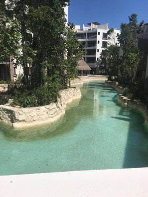 A clear water feature with a stone border, surrounded by greenery and modern buildings in the background. - Departamento en Playa del Carmen. (Playa del Carmen)