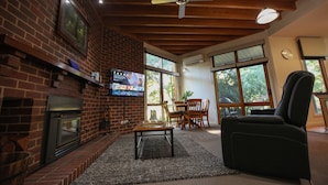 A living room with a brick fireplace, a television mounted on the wall, a ceiling fan, and large windows. - The Lodges (Halls Gap)