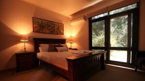 A bedroom with a large bed, two bedside tables with lamps, a painting on the wall, and a large window with a view of trees. - The Lodges (Halls Gap)