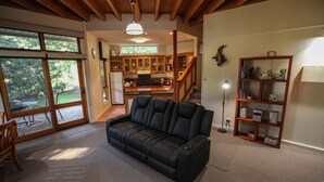 A spacious living room with a black leather sofa, wooden flooring, a large window, and a kitchen area visible through an open door. - The Lodges (Halls Gap)