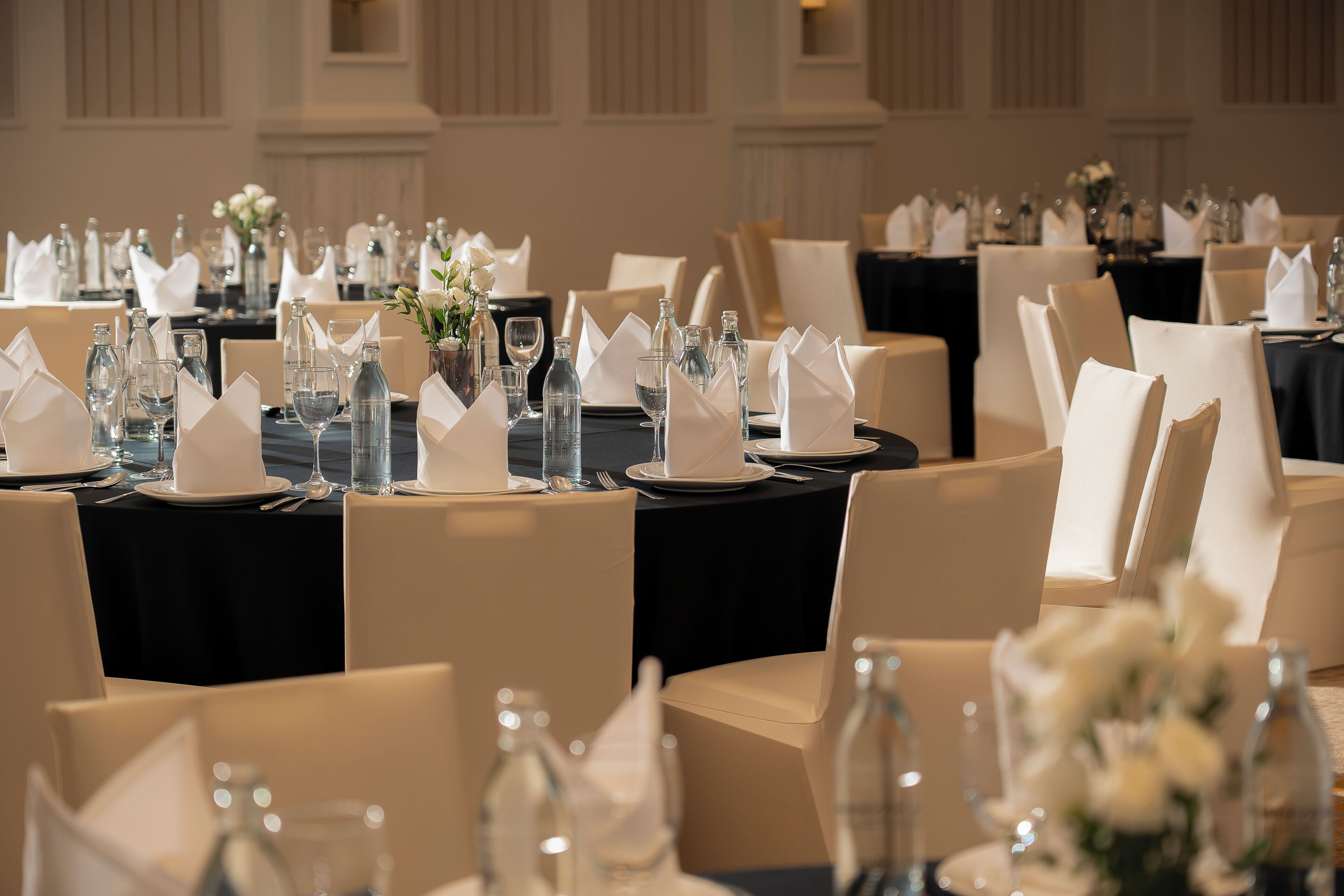 A banquet hall with round tables set for a formal event, featuring white tablecloths, glassware, and floral centerpieces.