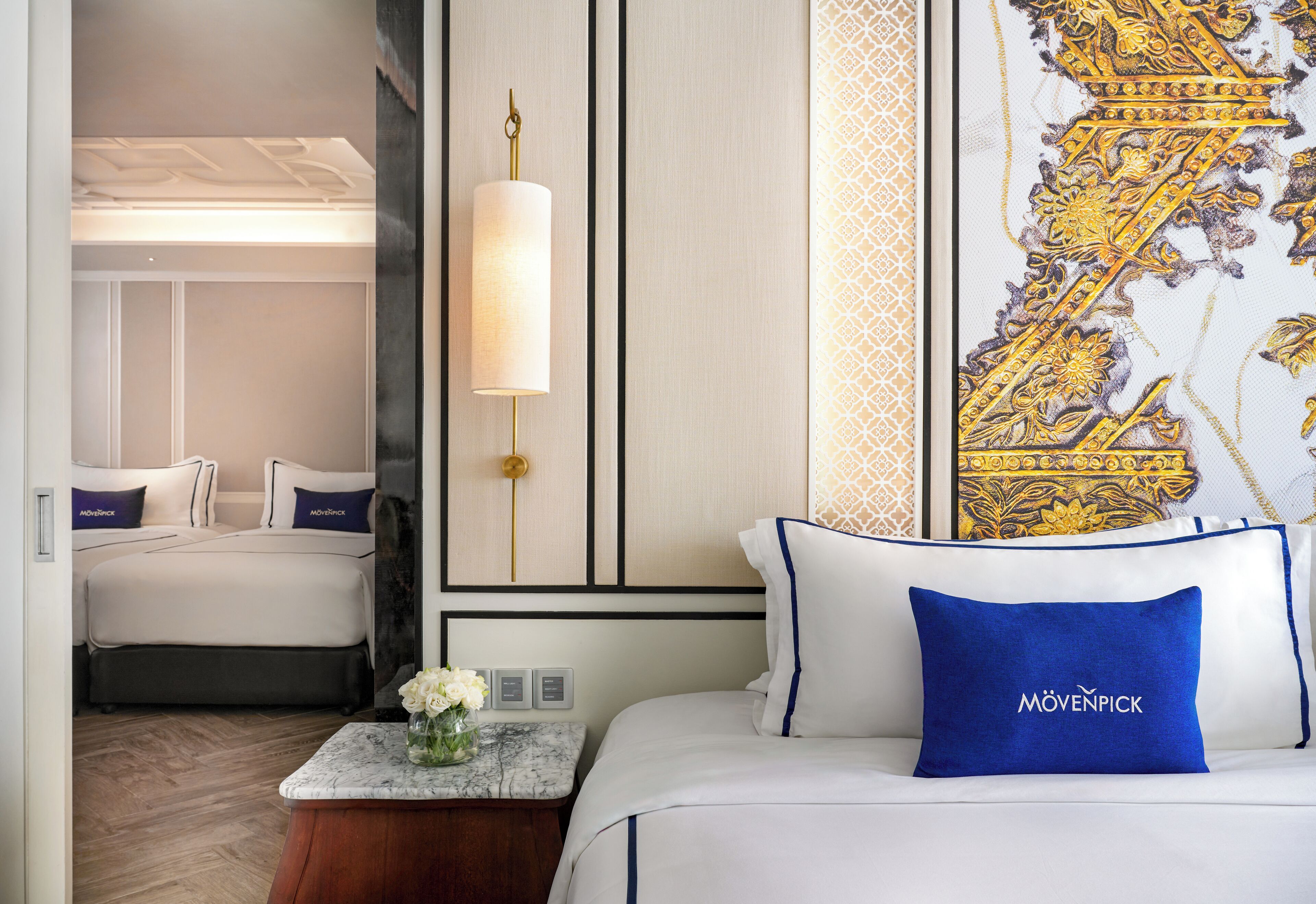 A hotel room with a bed, a nightstand with a vase of flowers, a wall-mounted lamp, and a decorative tapestry.