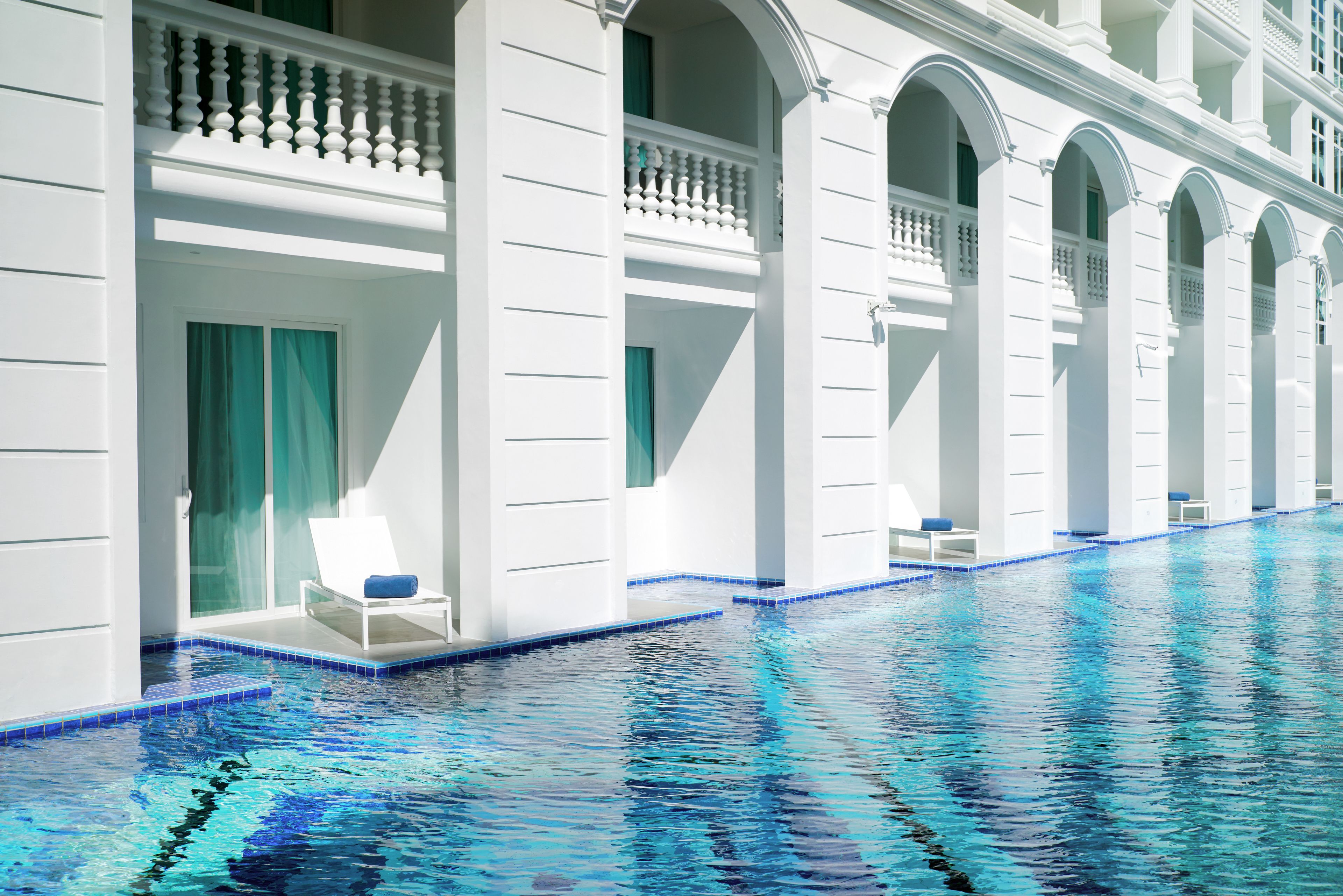 Una piscina con columnas blancas y bordes con azulejos azules, rodeada de muros blancos y balcones con cojines azules.