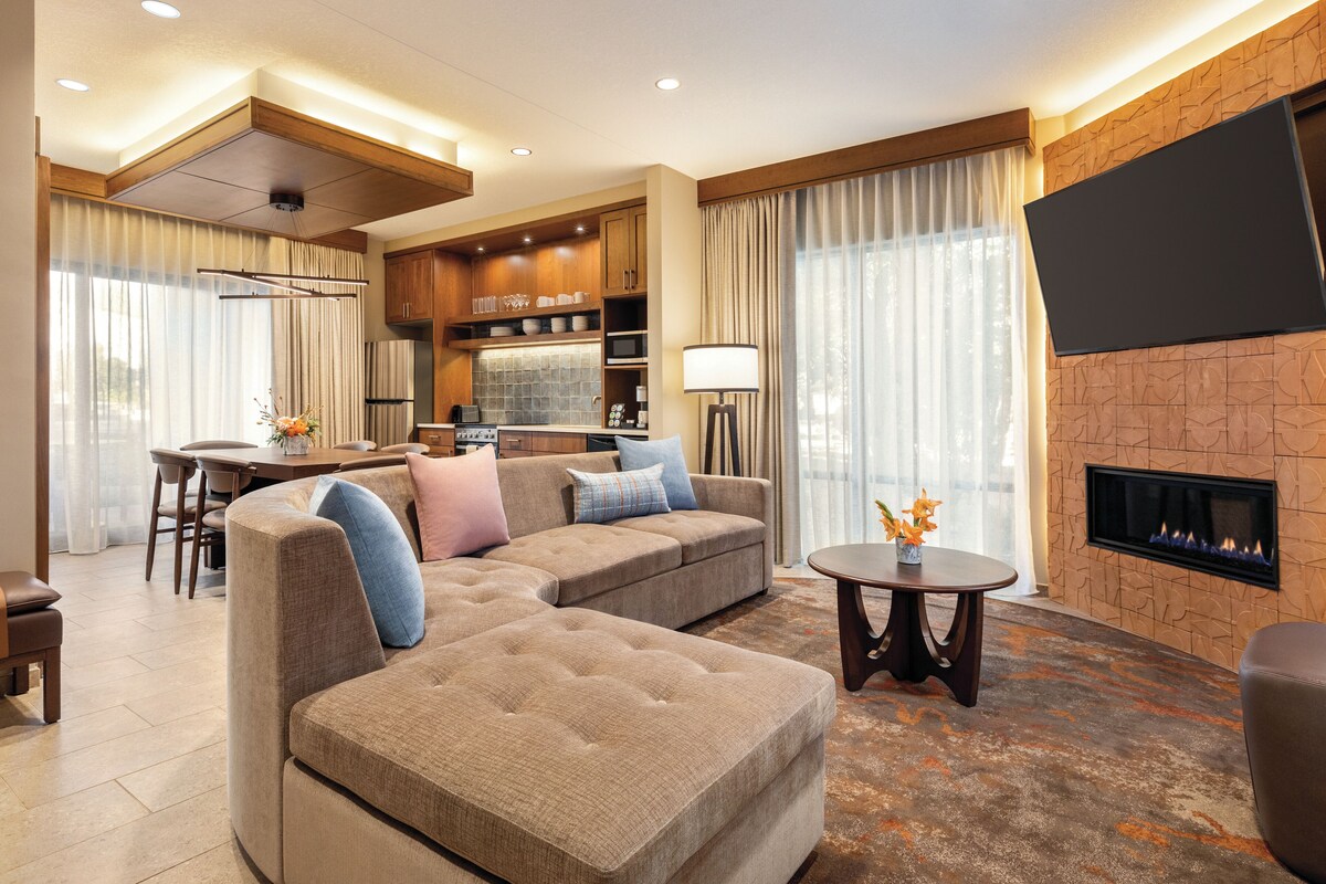 A modern living room with a sectional sofa, a fireplace, a flat-screen TV, and a kitchen area in the background.
