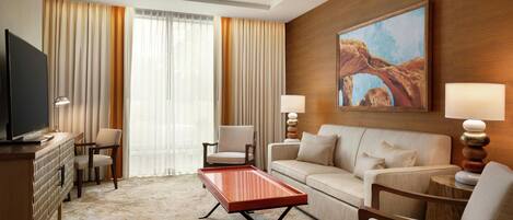 A hotel room with a flat-screen TV, a sofa, a coffee table, two armchairs, a dining table, and a painting on the wall.