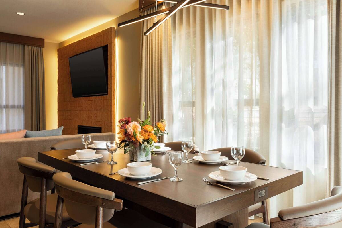 Un comedor moderno con una mesa de madera dispuesta para cuatro, un televisor de pantalla plana montado en una pared de ladrillo y un sofá al fondo.