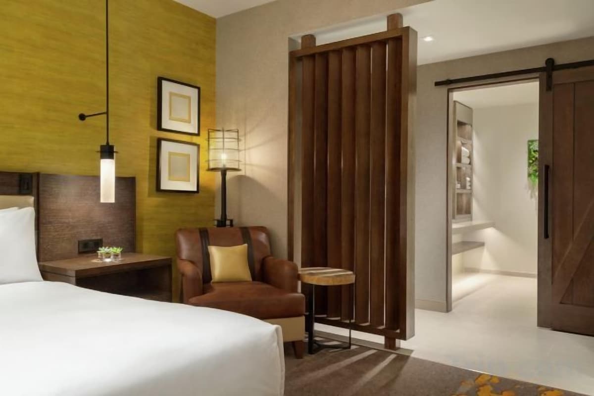 Een hotelkamer met een bed, een stoel, een klein tafeltje, een lamp en een schuifdeur naar een badkamer.