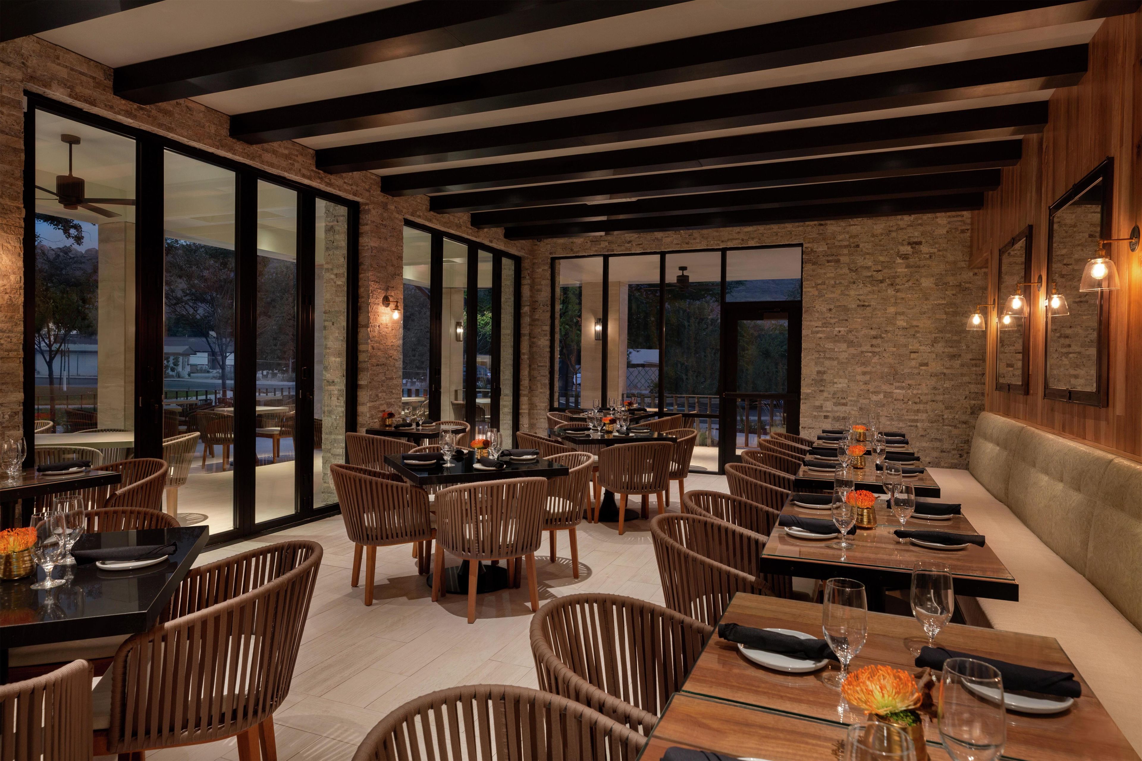 A restaurant with large windows, wooden ceiling beams, and wicker chairs. Tables are set with plates, glasses, and flowers.