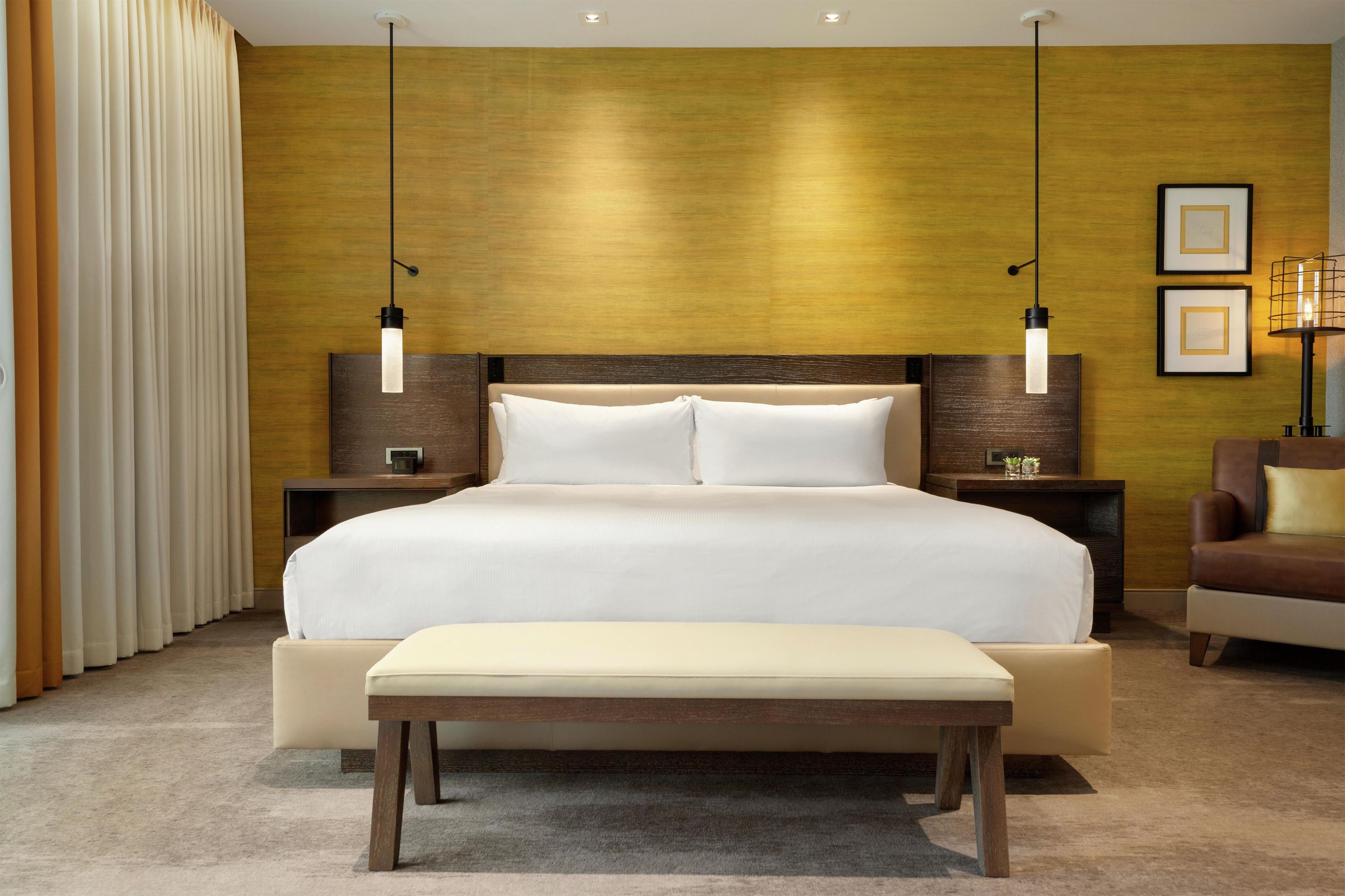 A hotel room with a large bed, bedside tables, a bench, a chair, and a wall with a textured finish.