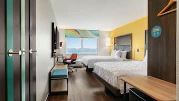 A hotel room with two beds, a desk, a chair, a bench, a TV, and a view of the sea.