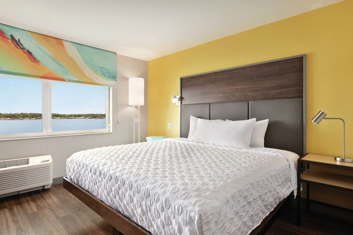 A hotel room with a large bed, a view of the water, a painting, and a bedside table with a lamp.