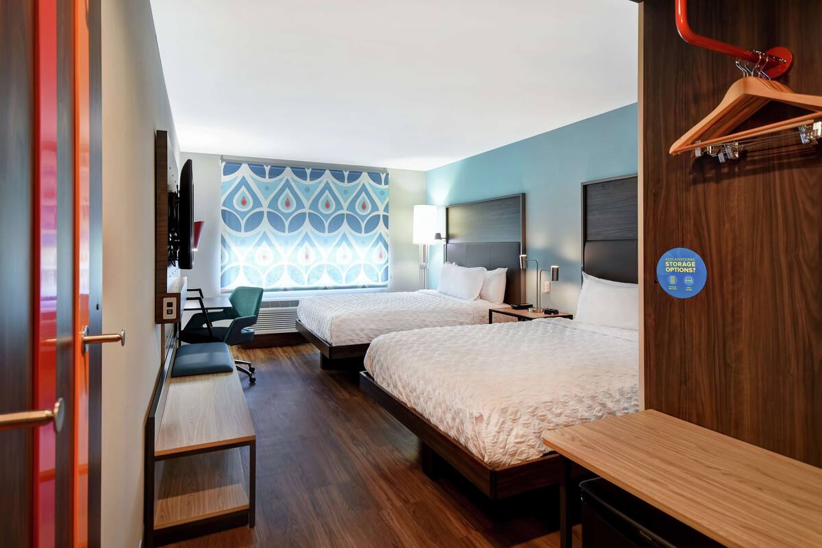 A hotel room with two beds, a desk, a chair, a TV, and a window with a patterned curtain.