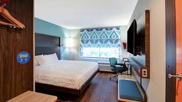 A hotel room with a bed, a desk, a chair, a TV, and a window with a geometric pattern.