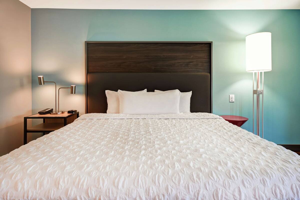 A neatly made bed with a textured white duvet, a dark headboard, a bedside lamp, and a modern floor lamp.