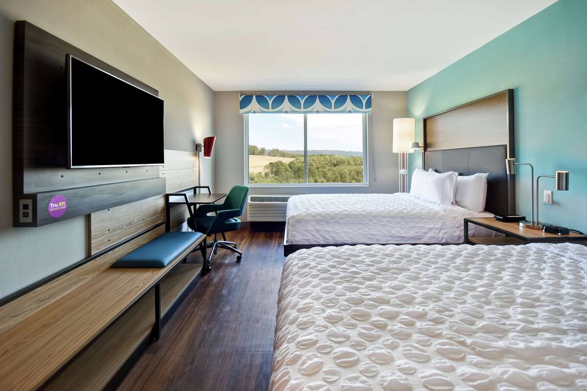 A hotel room with two beds, a desk with a chair, a TV, and a large window with a view of the countryside.